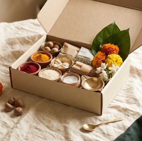 An open kraft box holding bowls of turmeric, kumkum and rice, cotton wicks, a brass lamp and flowers.
